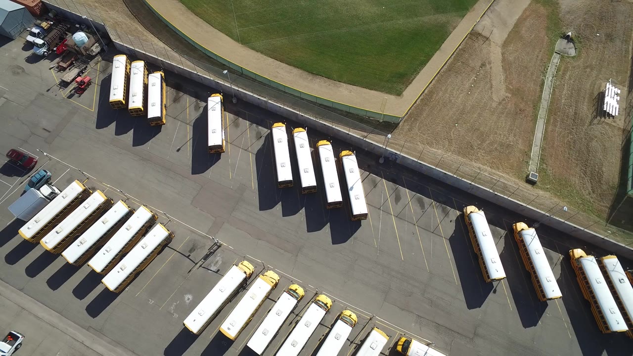 Aerial frame in Hidden Lake, Colorado showing rows of white and yellow buses, trucks, cars, pavement, dry grass, and a green field with no visible people.