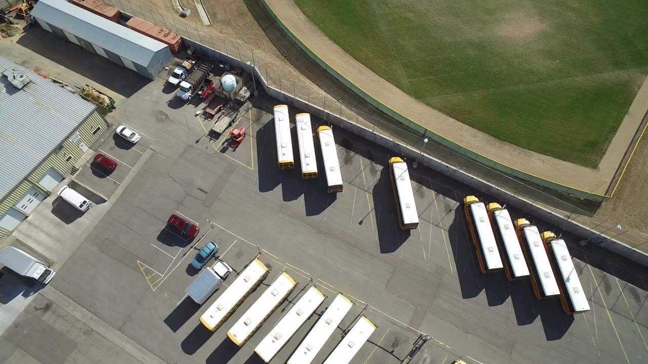 Overhead school bus parking lot beside a metal-roofed building and green field.