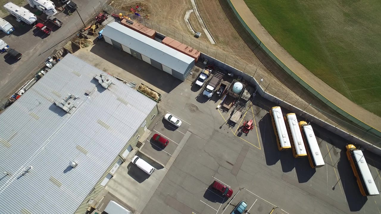 A high aerial frame in Hidden Lake, Colorado shows the industrial building roof, several parked white vehicles, long trailers or buses, a service yard, and a curved green field boundary with no visible people.