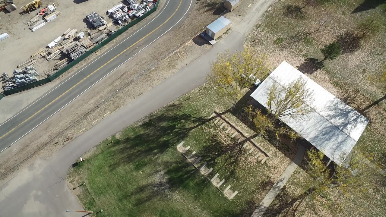High-angle aerial frame in Hidden Lake, Colorado showing a curved two-lane road, fenced industrial storage area, trees, lawn, and a white-roofed building, with no visible people.