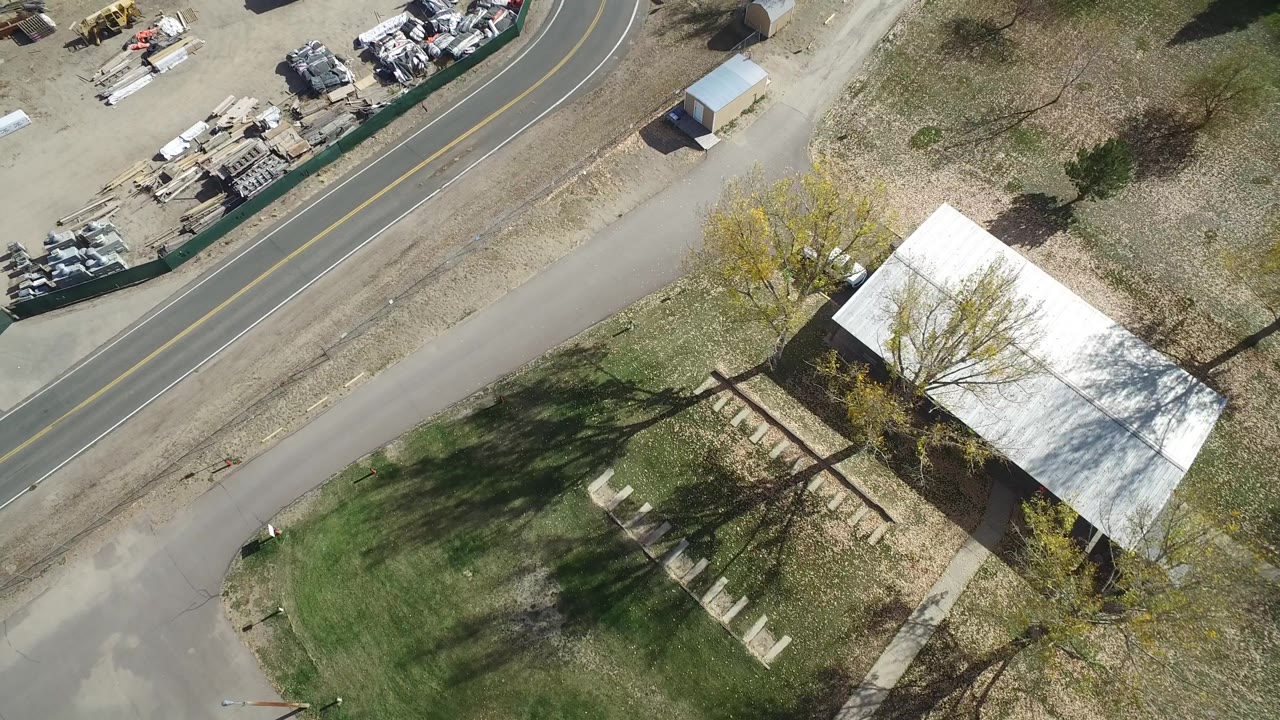 Aerial frame from Hidden Lake, Colorado with a white car on the curved road, stacked materials in a fenced yard, dry grass, trees, and the roof of a nearby building, with no visible people.