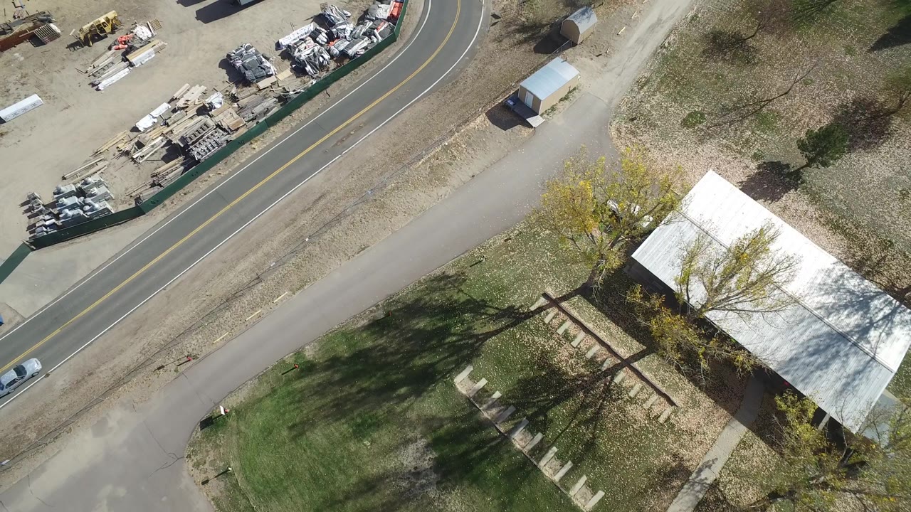 Hidden Lake, Colorado aerial frame showing a white car farther along the curved two-lane road, a gravel access lane, fenced storage materials, trees, and dry open ground, with no visible people.