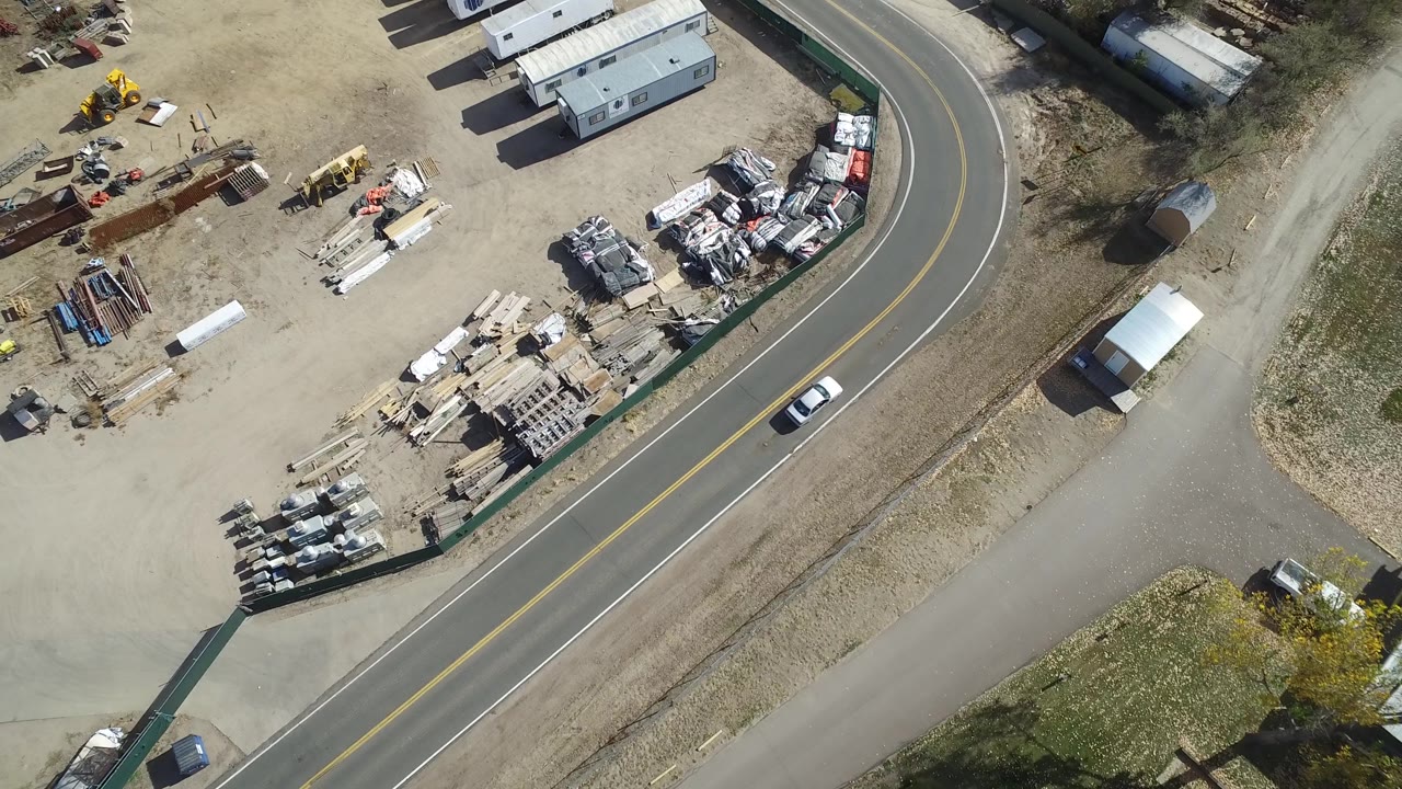A high overhead view in Hidden Lake, Colorado shows the white vehicle farther along the curved road beside the dirt yard, trailers, equipment piles, and no visible people.