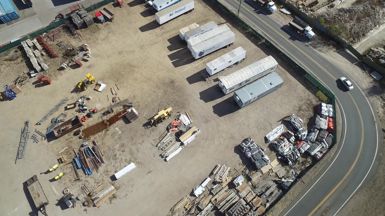Overhead view of trailers, work materials, equipment, and a curved road beside a dusty lot.