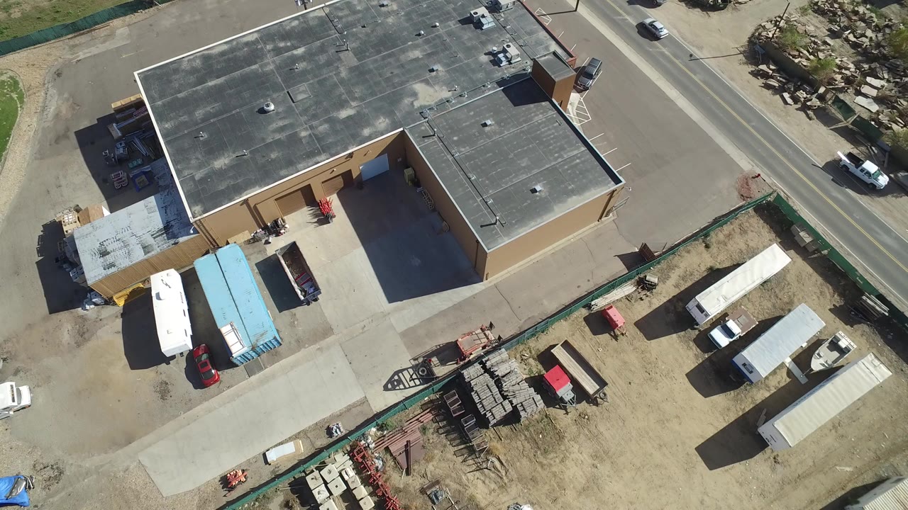 Overhead view of a tan flat-roof building beside a paved road and work yard.