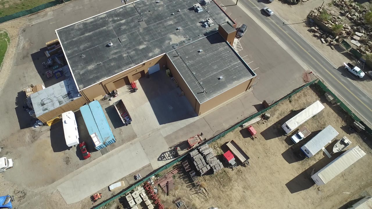 Drone-height view in Hidden Lake, Colorado with the flat roof centered, a road along the upper right, white trailers and blue-roofed storage at lower left, and no visible people.