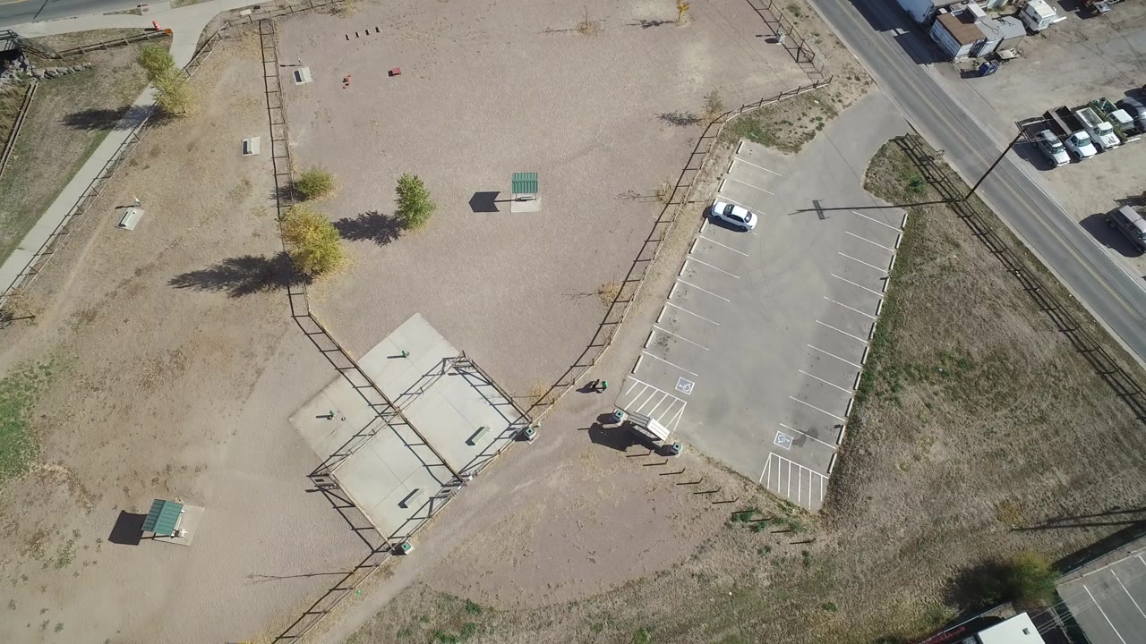 High overhead frame in Hidden Lake, Colorado showing a marked parking lot, dry vacant ground, a roadside edge, scattered small trees, and no visible people.