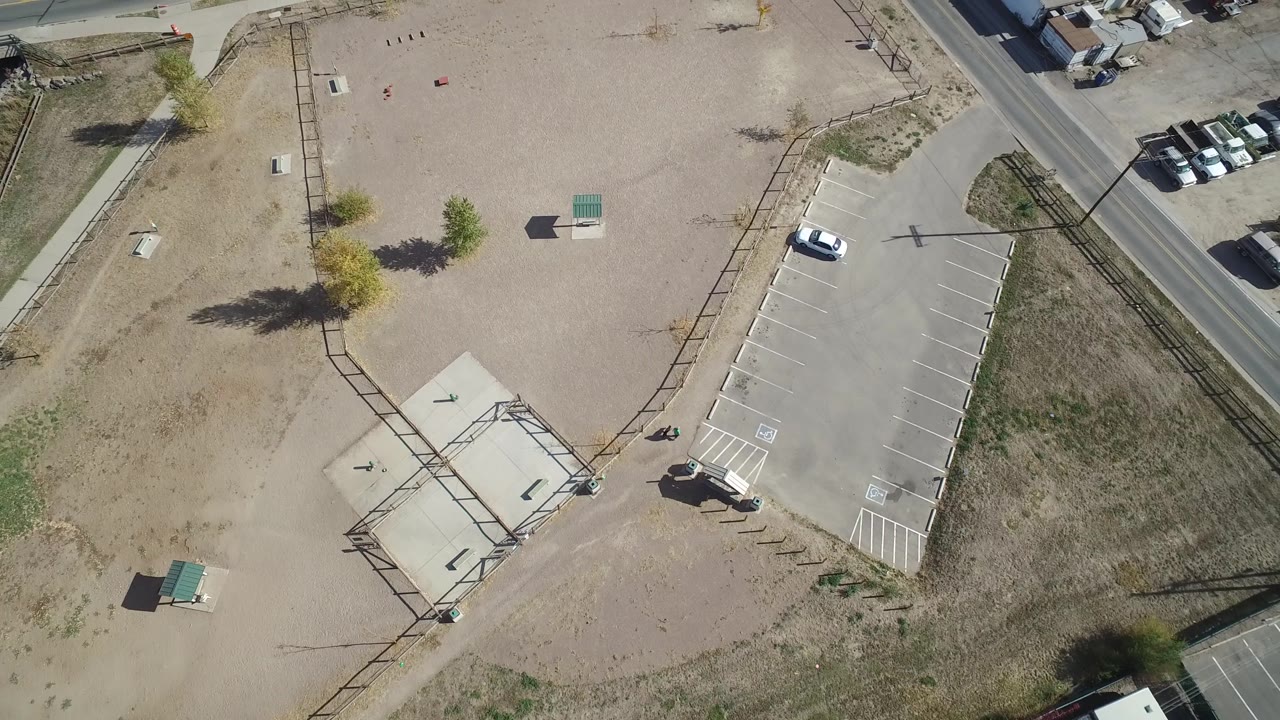 Drone-like overhead view in Hidden Lake, Colorado with the parking area shifted toward the right, dry vacant land across the center, roadside pavement above, and no visible people.