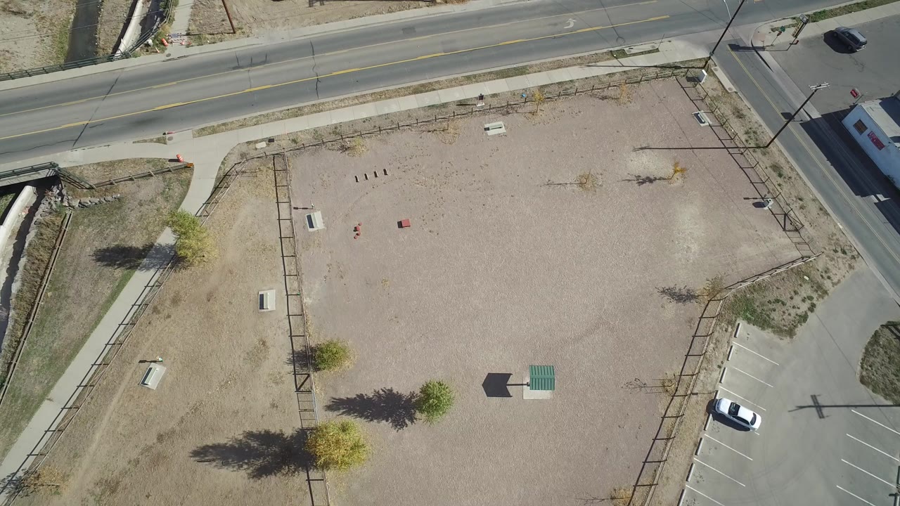 Overhead drone view in Hidden Lake, Colorado showing a broad gravel lot, paved road, sidewalks, parking spaces, small trees, and no visible people.