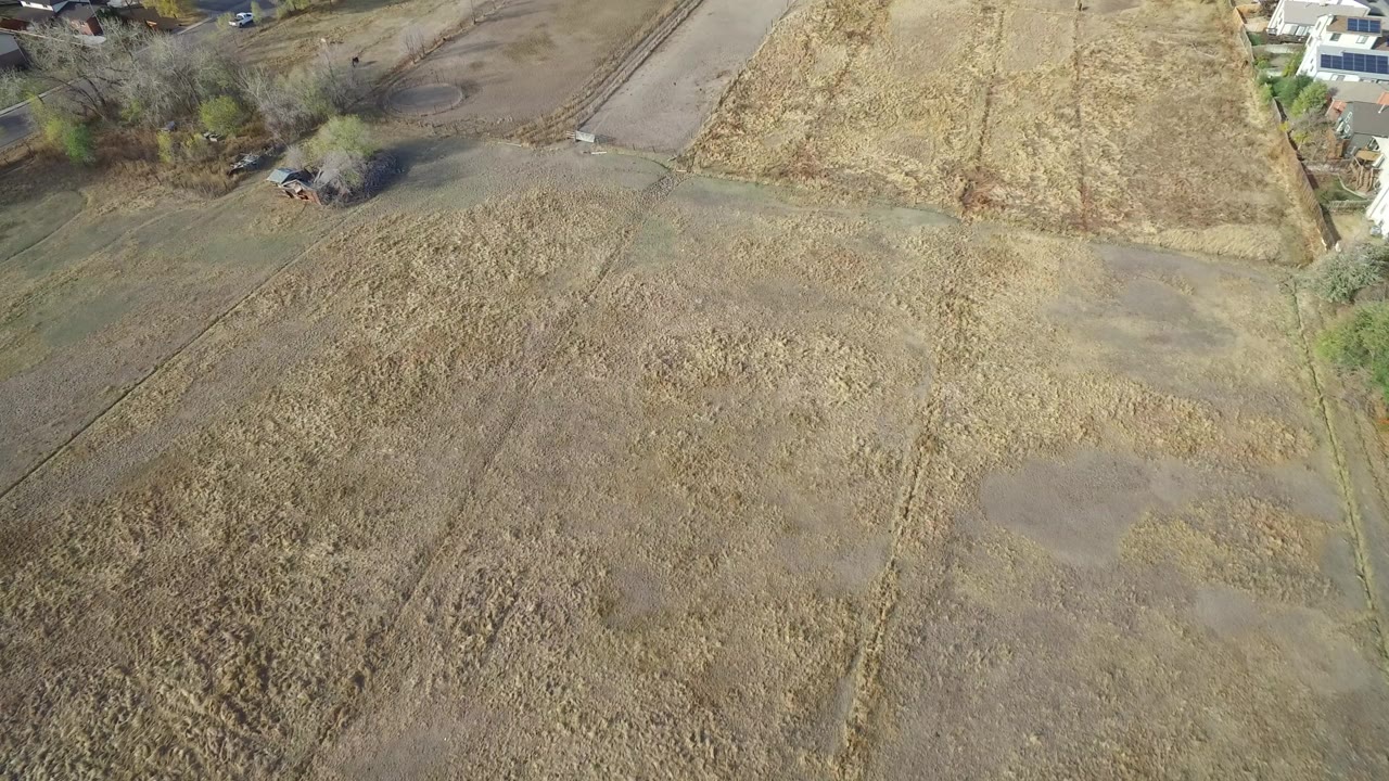High-angle drone frame over dry open terrain in Hidden Lake, Colorado, with faint tracks, a road edge, scattered trees, partial homes at the top edge, and no visible people.