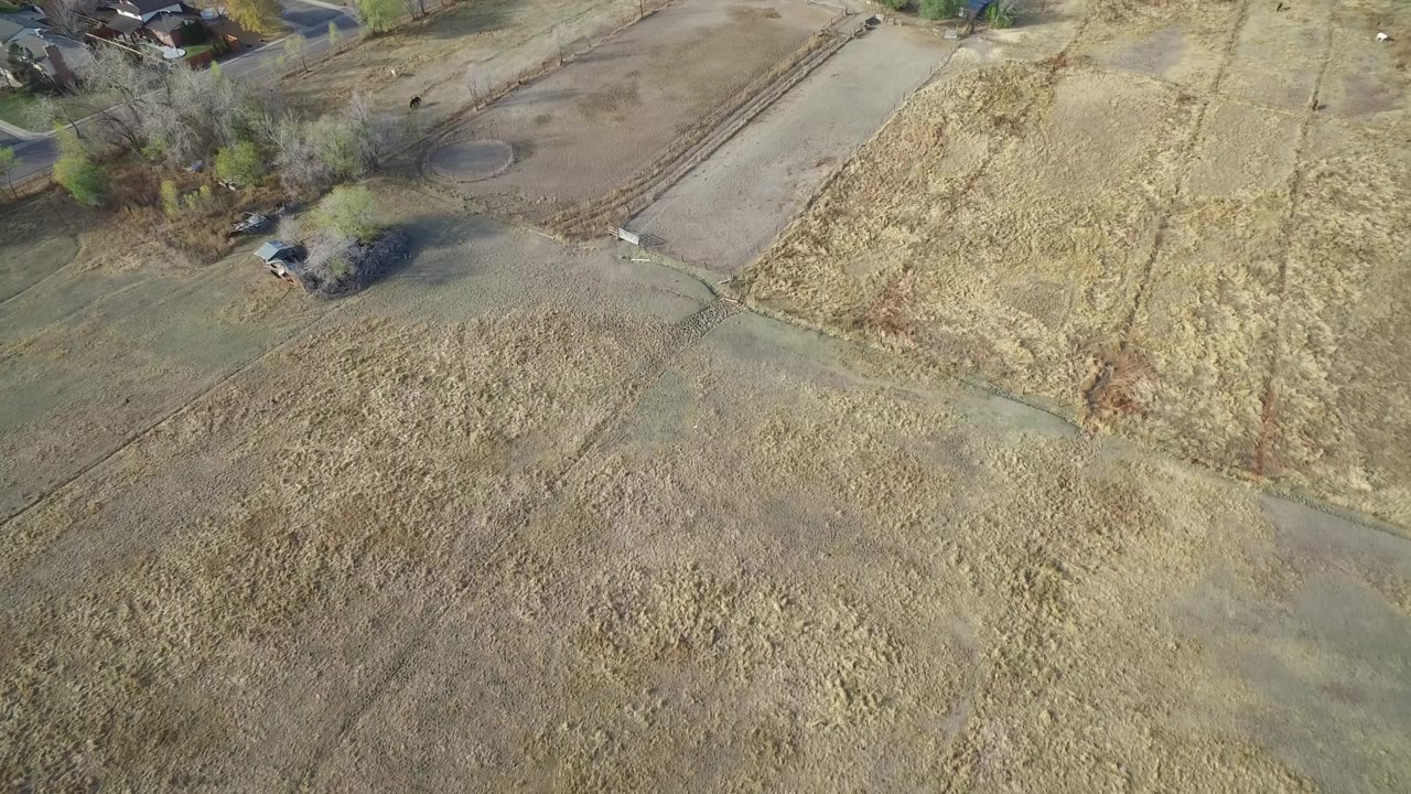 Drone-style view of dry fields divided by tracks with houses and vehicles above.