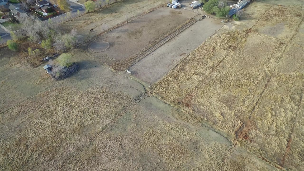 Aerial frame over Hidden Lake, Colorado with pale grassland in the foreground, a dirt lot and fenced lines in the midground, homes and vehicles near the top, and no visible people.