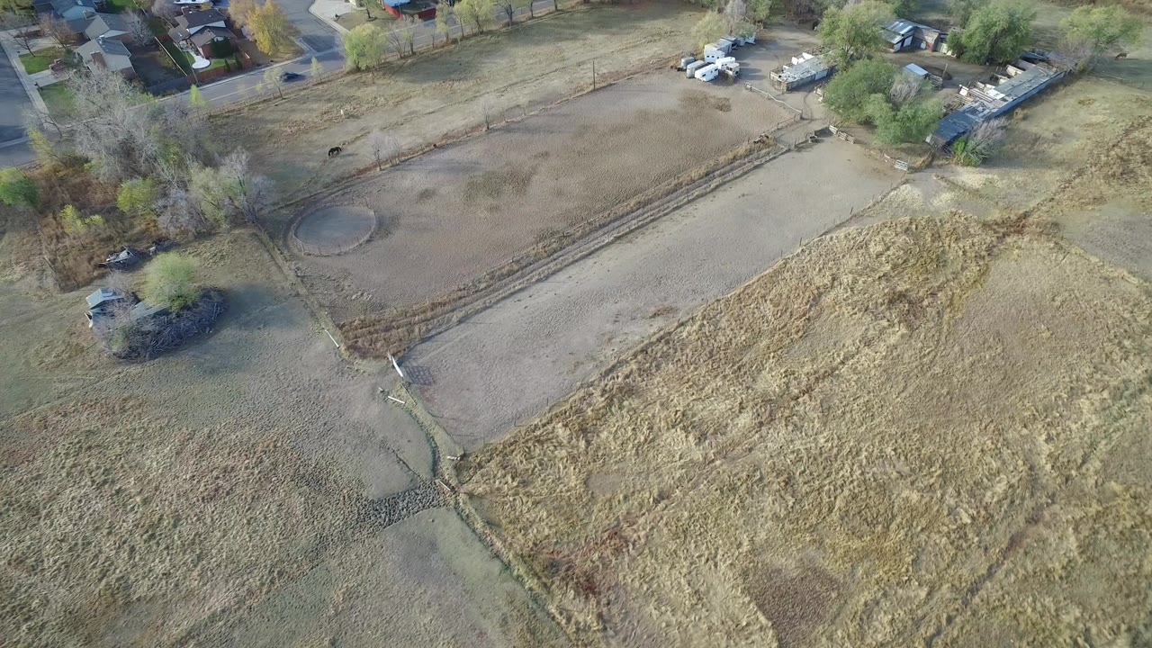 Aerial view of dry fields, trees, houses, and parked vehicles.