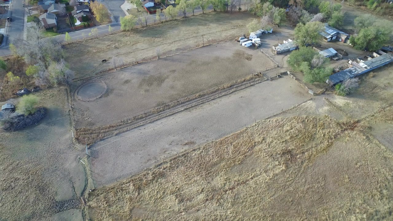 Aerial view of dry fields, a dirt plot, parked vehicles, trees, and nearby homes.