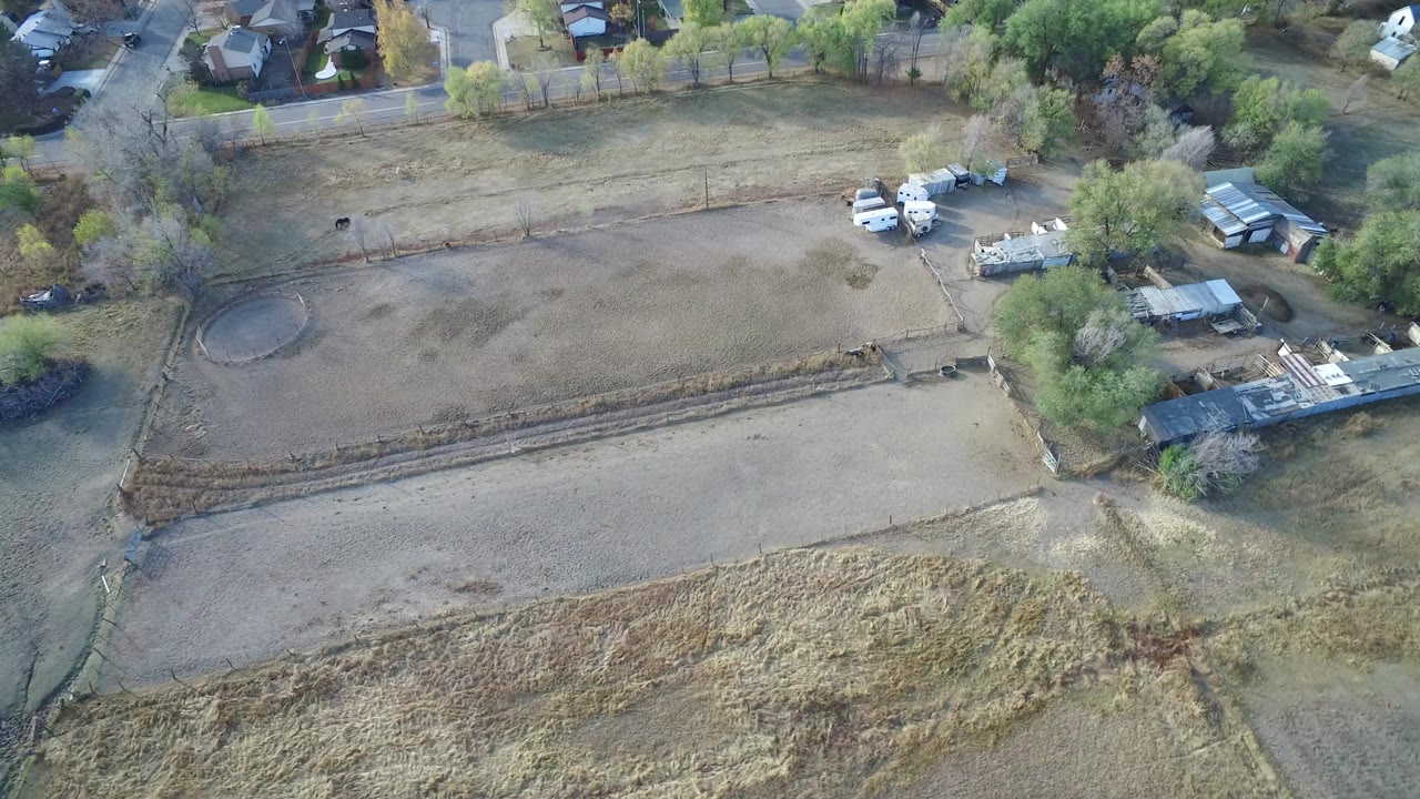 Opening aerial frame over vacant light brown lots in Hidden Lake, Colorado, with trees, small buildings, parked white vehicles, nearby homes, and no visible people.