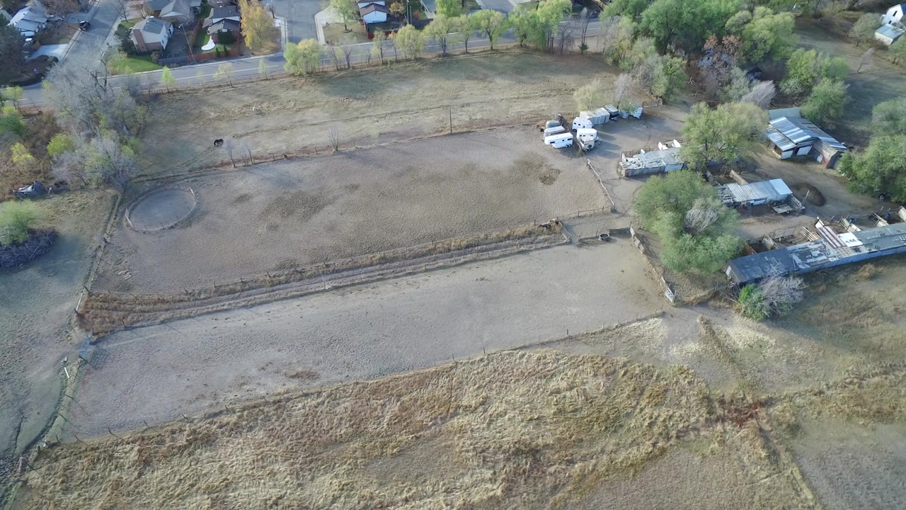Aerial frame in Hidden Lake, Colorado showing dry vacant parcels, a circular ground feature, trees, small structures, parked vehicles, homes at the upper edge, and no visible people.