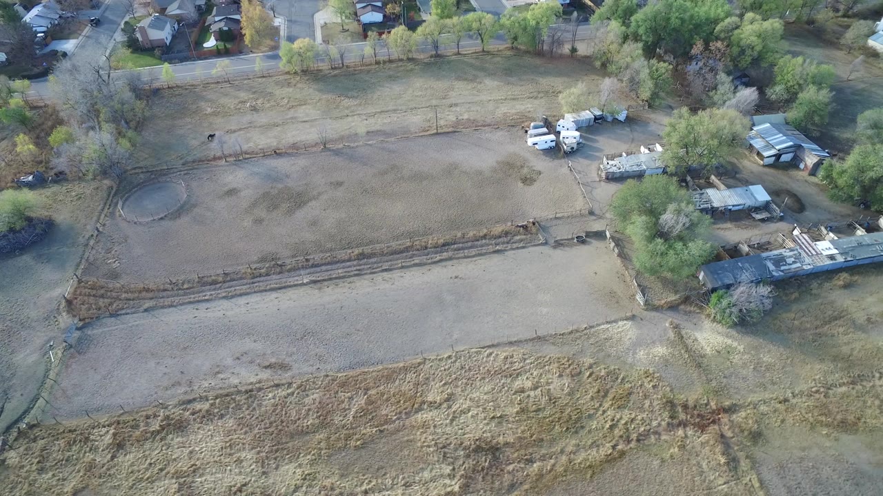 Drone view of open lots, fenced edges, parked vehicles, sheds, trees, and residential roofs.