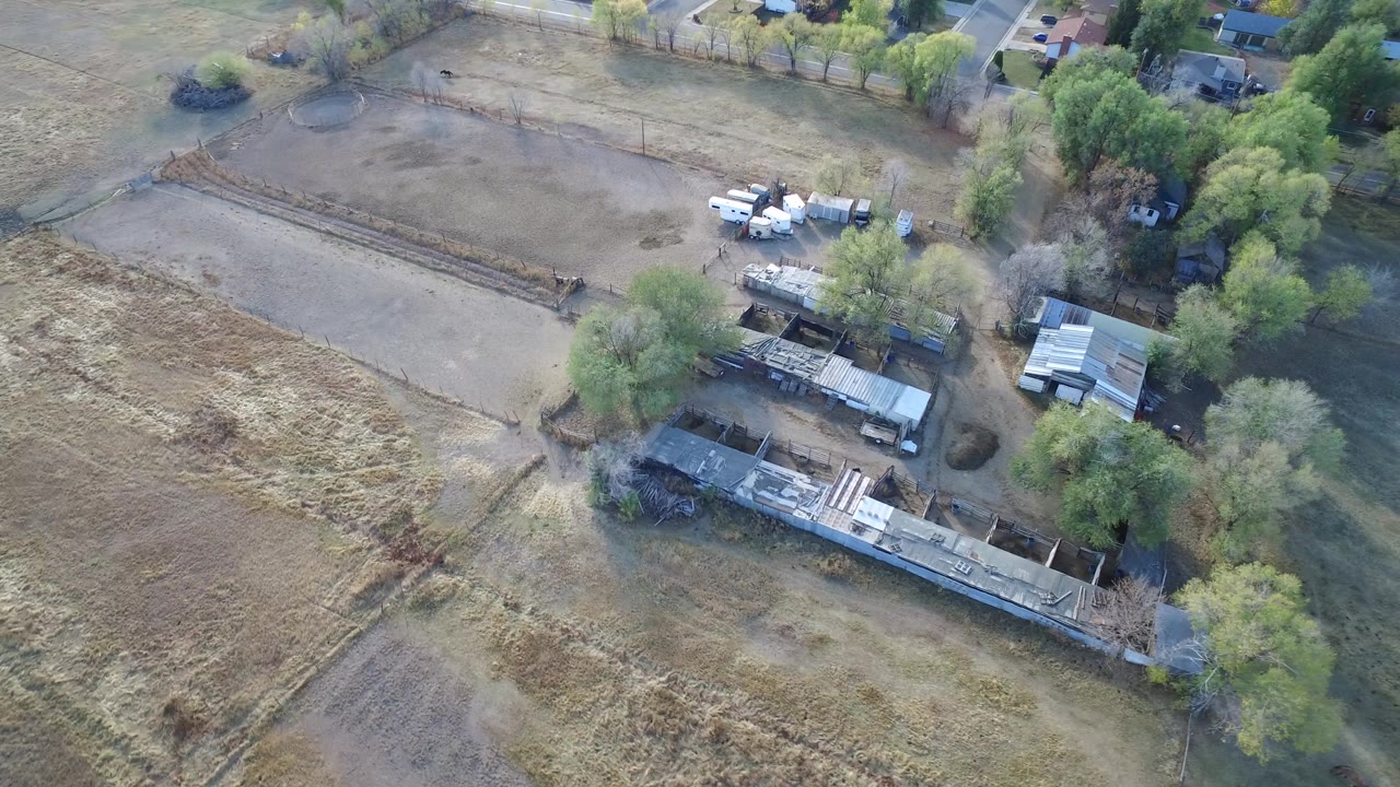 Aerial view of dry fields, trees, small buildings, and nearby homes.
