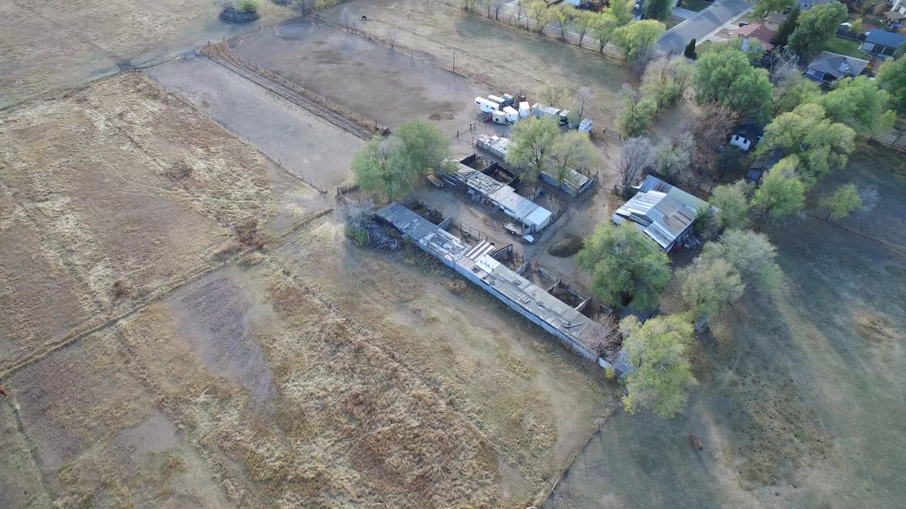 High aerial frame over Hidden Lake, Colorado showing dry grassy fields beside a long low building, scattered trees, nearby homes, and no visible people.