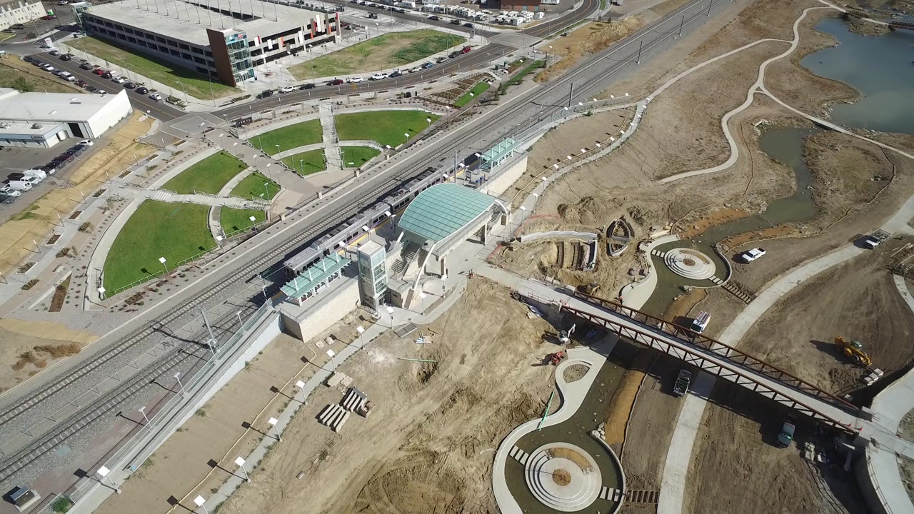 High aerial view in Hidden Lake, Colorado showing rail tracks, a glass-roofed station, roads, green lawns, ponds, and bare construction ground, with no visible people.