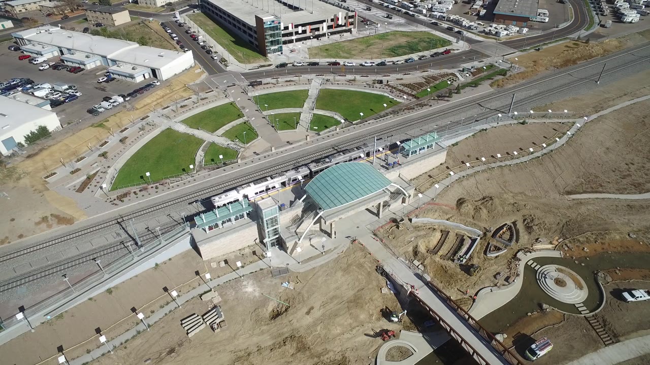 Aerial view in Hidden Lake, Colorado with the rail line running across the frame, a teal station roof near the lower center, curved walkways, small green lawns, parked vehicles, and no visible people.