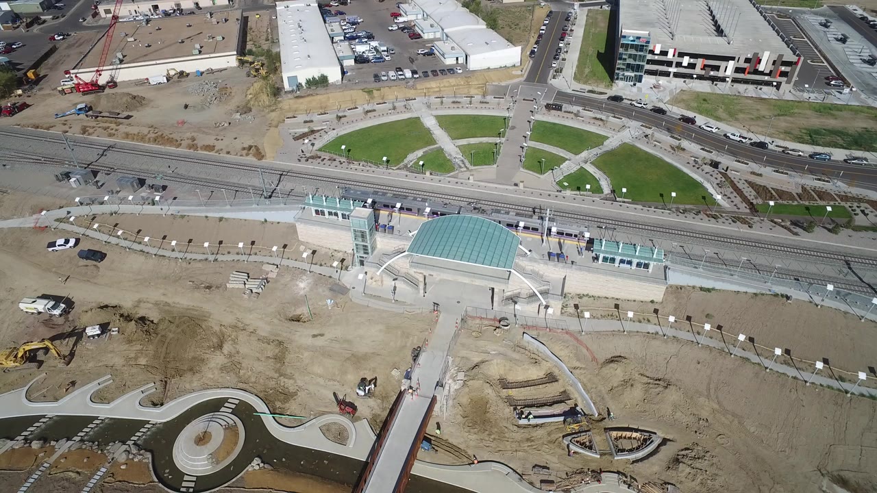 High-angle aerial frame over Hidden Lake, Colorado, showing rail tracks, a teal-roofed commercial building, paved roads, graded dirt, and no visible people.