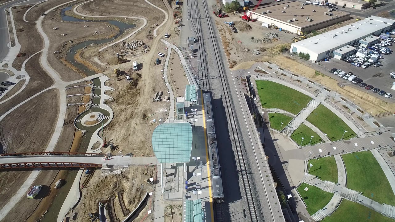 High aerial frame over Hidden Lake, Colorado showing rail tracks beside a road, a red pedestrian bridge, green lawns, brown fields, parking areas, industrial buildings, and no visible people.