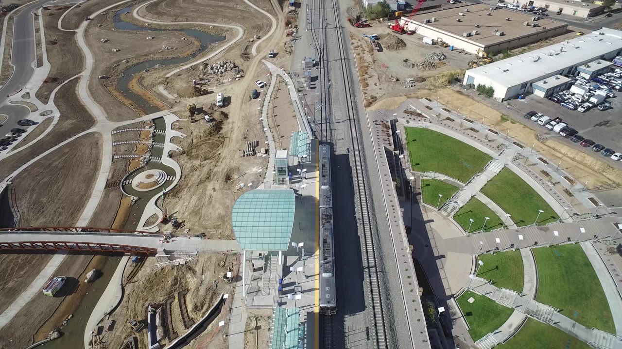Top-down aerial of transit tracks, paths, lawns, fields, and an industrial parking lot.