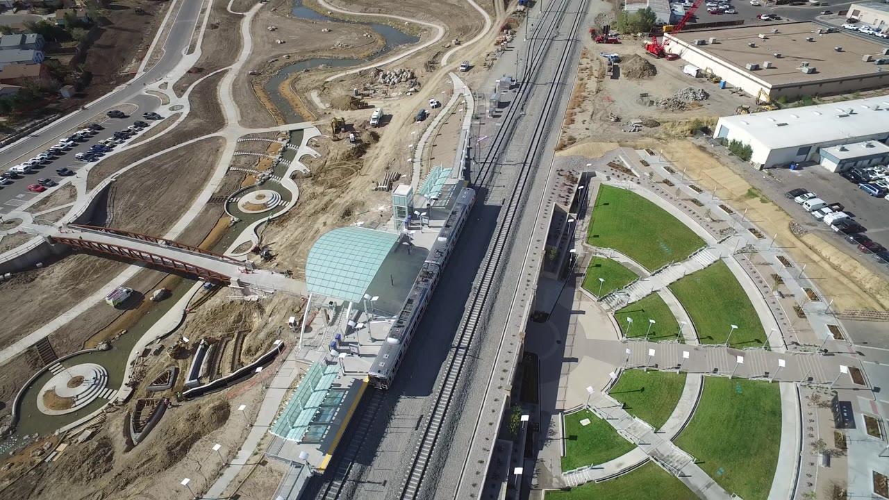 Opening aerial frame over a Hidden Lake, Colorado rail station area with tracks, a pale curved station roof, a pedestrian bridge, green lawn panels, roads, and no visible people.