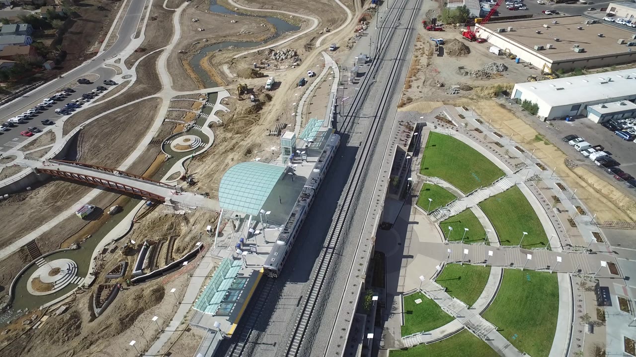 Aerial frame in Hidden Lake, Colorado showing the station roof closer to the lower right, rail lines running vertically, a rust-colored pedestrian bridge, circular landscaping, and no visible people.