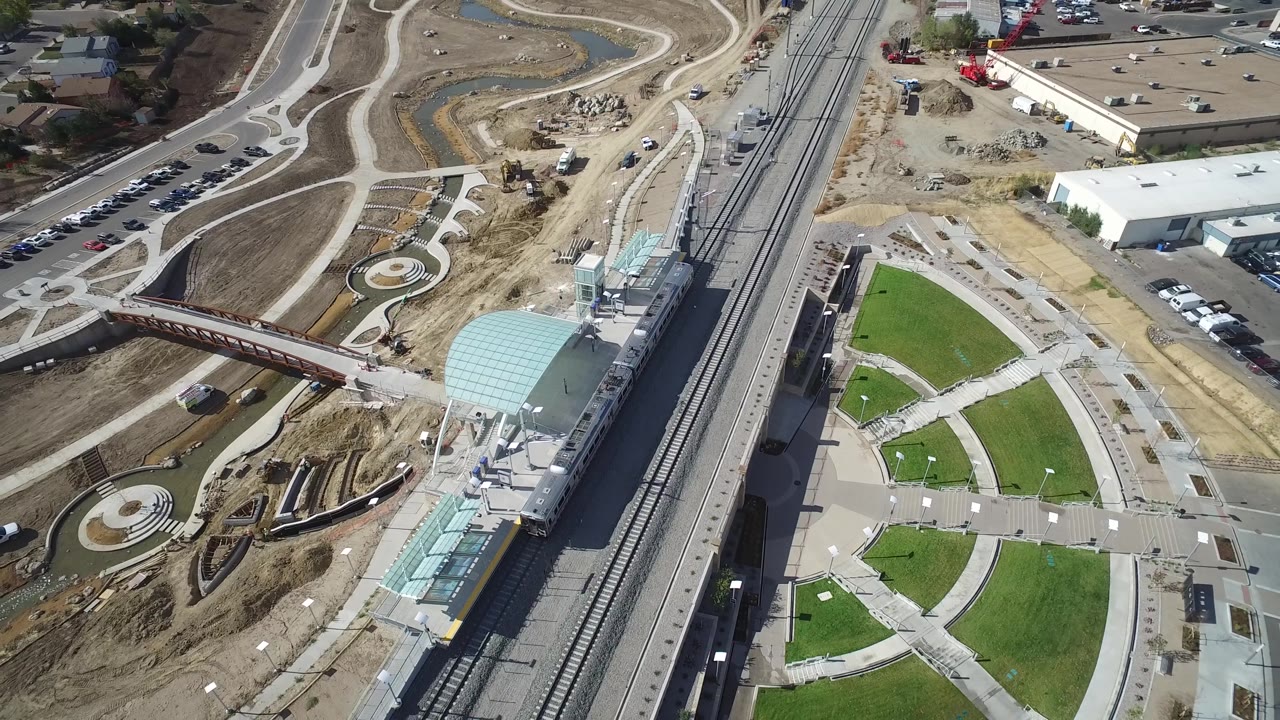 Drone-style view of station tracks, bridge crossing, green park lawns, white industrial building, and construction areas.