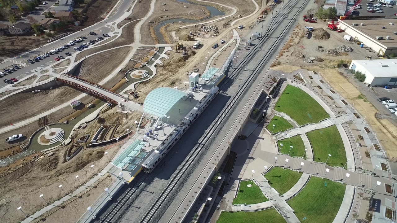 Opening aerial frame over Hidden Lake, Colorado shows rail tracks beside a station canopy, a pedestrian bridge, parking areas, road curves, landscaped paths, and bare construction ground with no visible people.