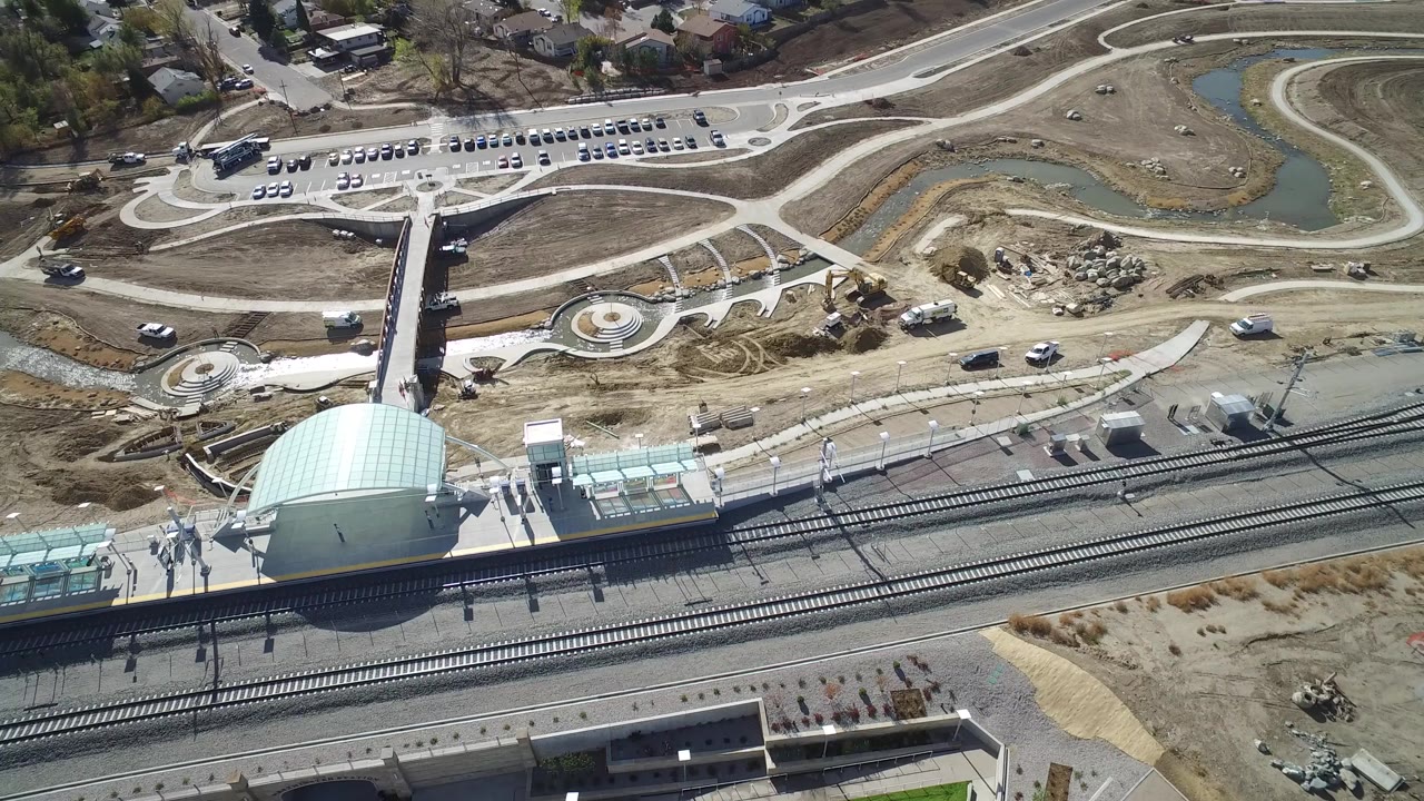 Aerial view of rail tracks, a station canopy, paths, ponds, parked cars, and bare ground.