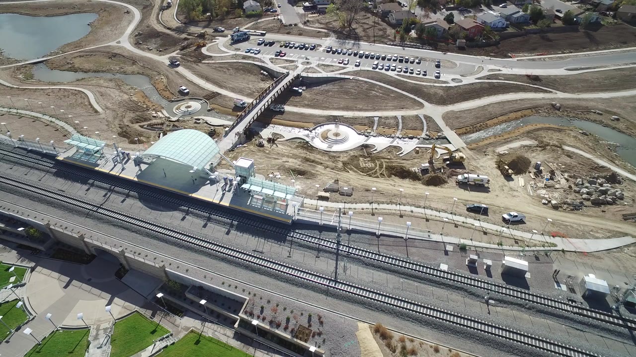 Aerial view of train station, rail tracks, pedestrian bridge, car park, ponds, and landscaping.