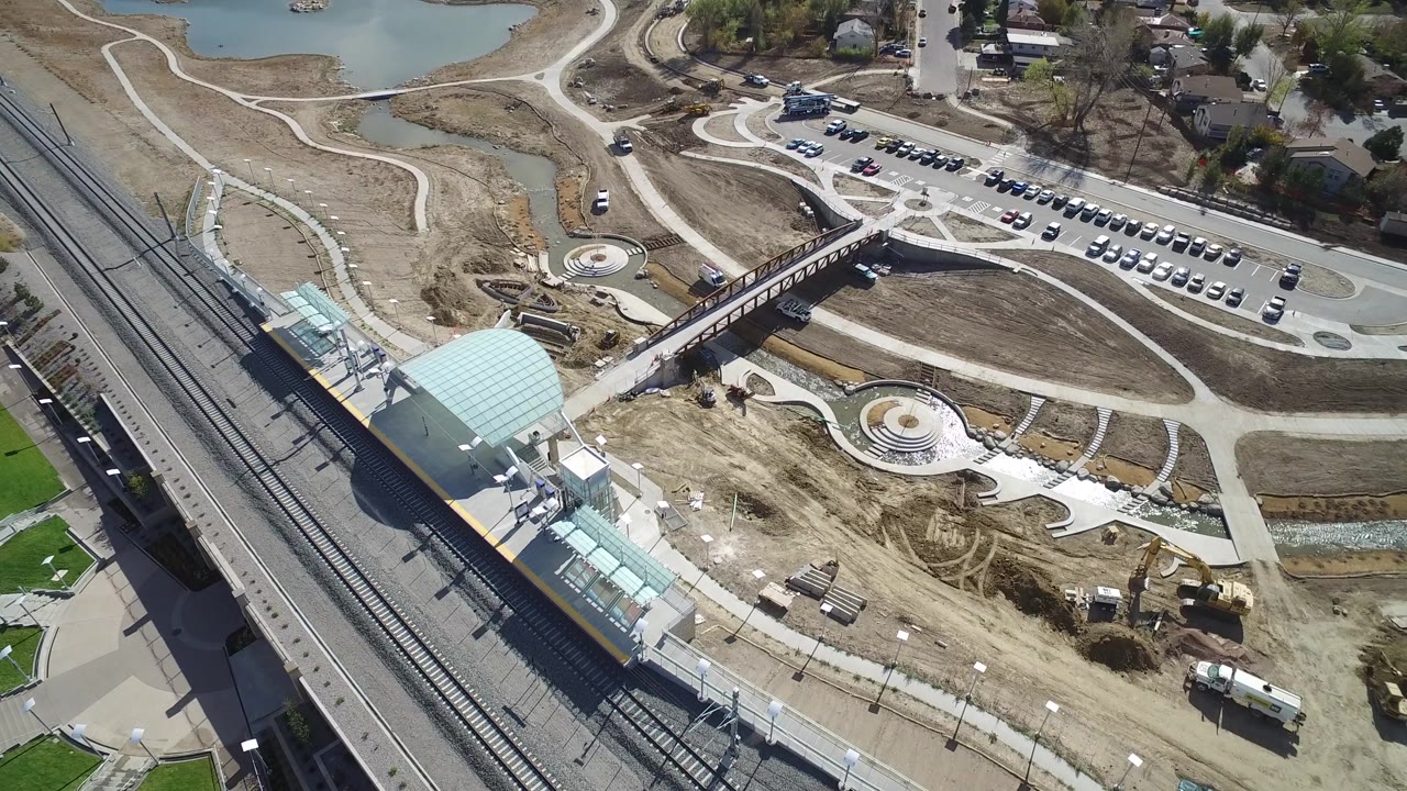 Opening aerial frame over Hidden Lake, Colorado showing rail tracks at left, a blue-roofed station structure, a pedestrian bridge, parked cars, curving paths, construction dirt, a pond edge, and no visible people.
