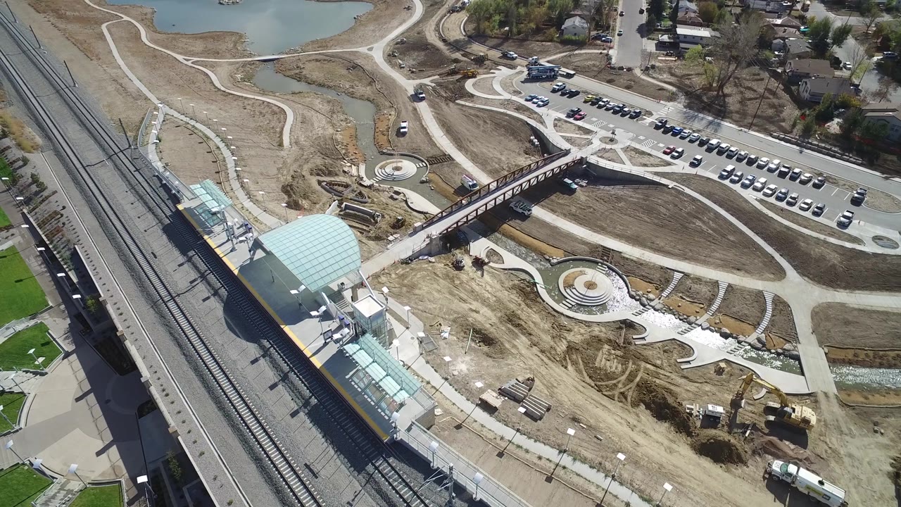 Bird's-eye view of rail tracks, station canopy, bridge, circular paved feature, parking rows, roads, pond, and graded dirt.
