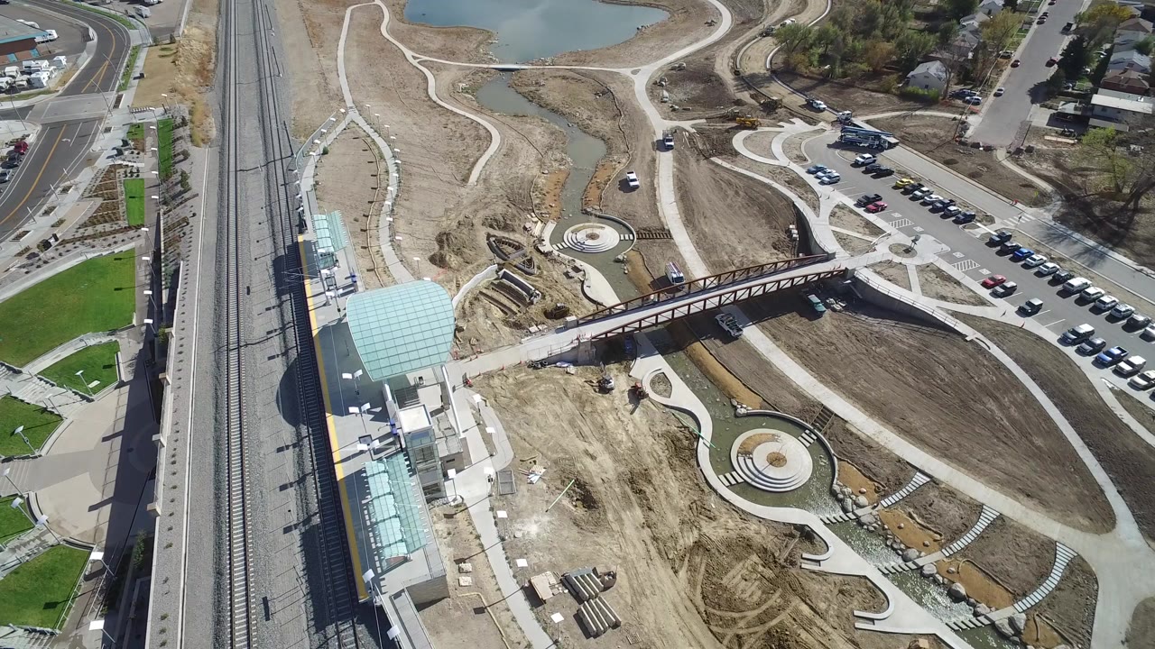 Aerial view of rail tracks, station roof, bridge, paths, parking, dirt lots, lawns, and water.
