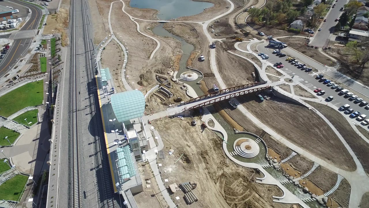 Aerial frame in Hidden Lake, Colorado with the pedestrian bridge centered, rail tracks running vertically at left, parking and road access to the right, green lawn patches, dirt construction areas, and no visible people.