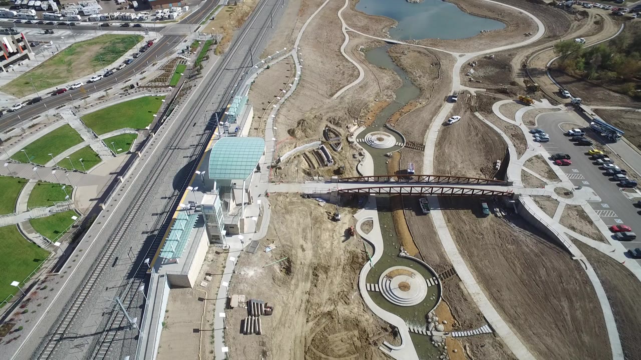 High aerial frame over Hidden Lake, Colorado showing rail tracks on the left, glass-roofed station structures, a pedestrian bridge, bare construction ground, curved paths, road traffic, and water at the top.