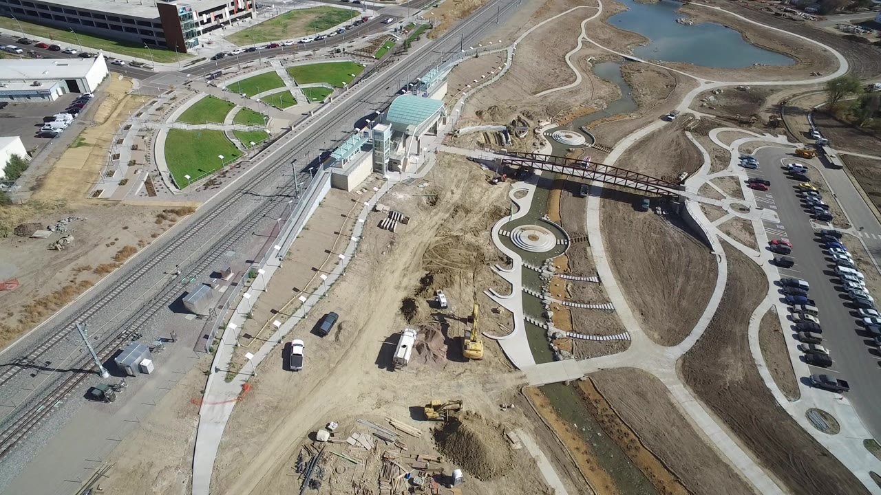 High aerial frame over Hidden Lake, Colorado showing diagonal rail tracks, curved pedestrian paths, a bridge, construction dirt, parked cars, ponds, and no visible people.