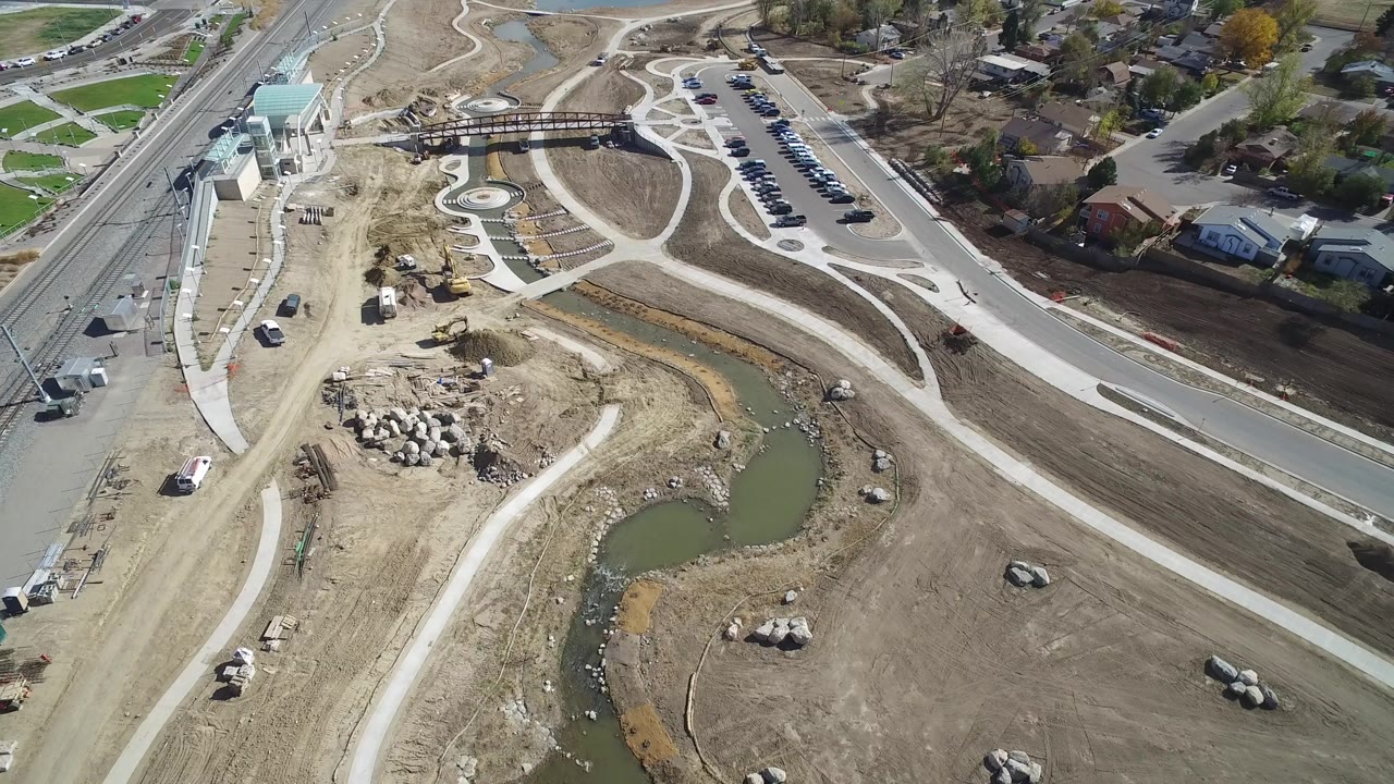 High aerial frame over Hidden Lake, Colorado, showing a green water channel, pale curved paths, a road, parked cars, construction ground, and nearby houses.