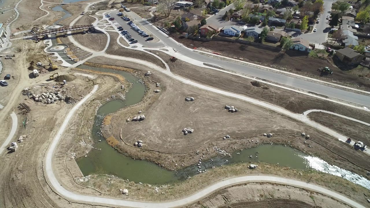 Drone-style aerial view in Hidden Lake, Colorado with a green water channel, open dirt area, roadside houses, small vehicles, and no visible people.