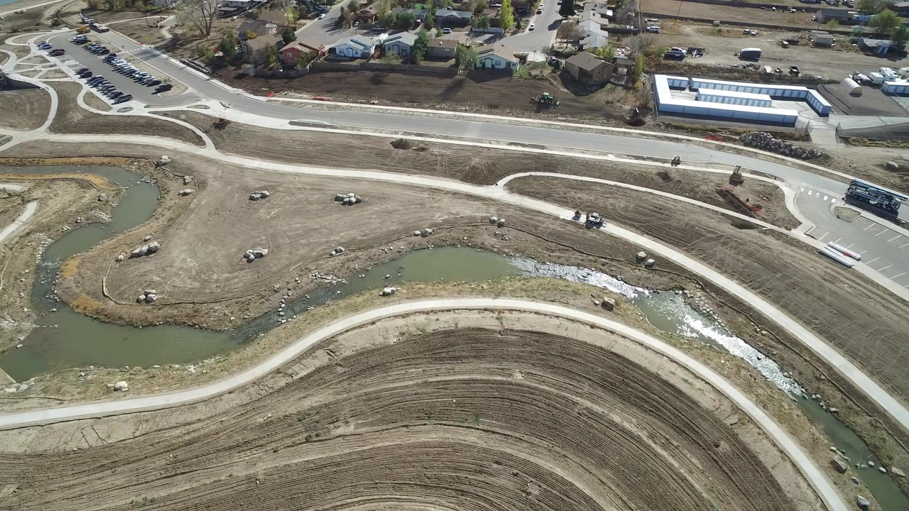 Aerial view of a canal, curved dirt grading, roads, parking, and nearby buildings.