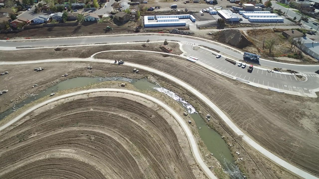 This Hidden Lake, Colorado frame keeps the sweeping brown ground pattern in the foreground with the drainage channel at left, a paved road at top, small vehicles, buildings, and no visible people.