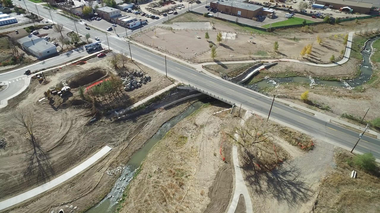 Aerial Hidden Lake, Colorado frame with the road angled across the foreground, a creek bend behind it, a small bridge or culvert, parked vehicles, and dry open ground with no clearly discernible people.