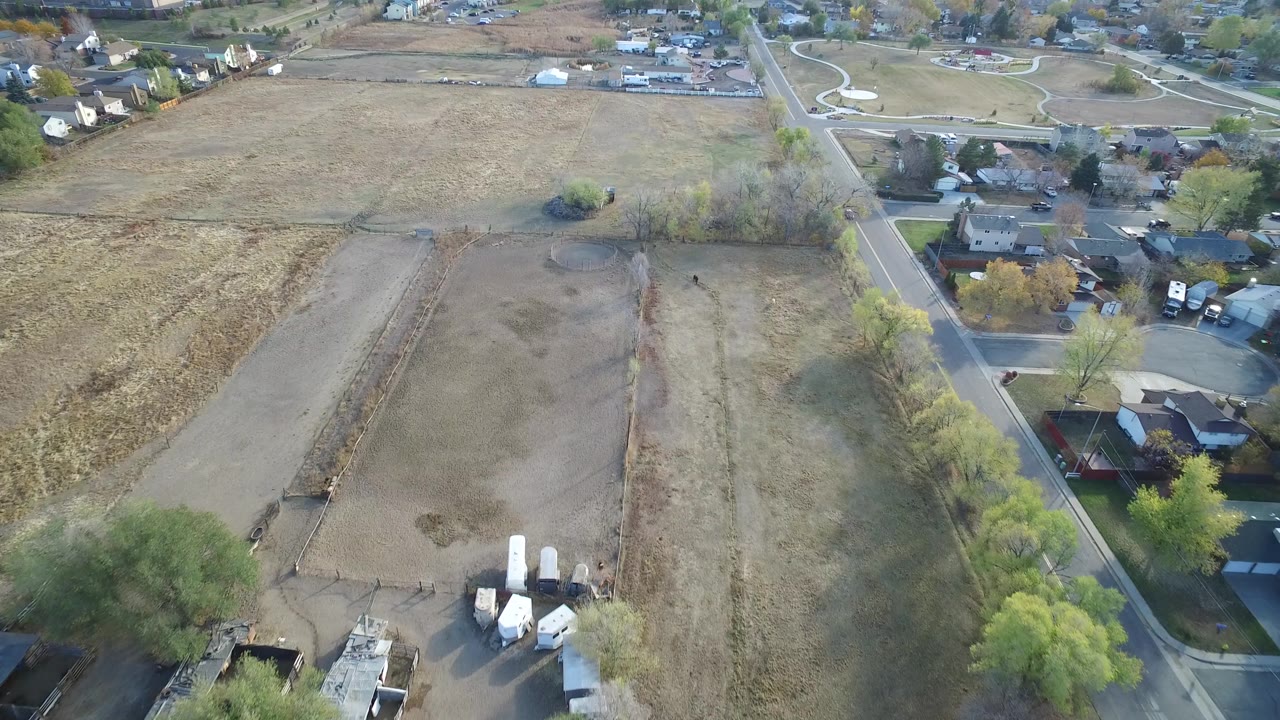 Aerial frame from Hidden Lake, Colorado showing an open brown field in the foreground, a road and homes to the right, scattered trees, and no visible people.
