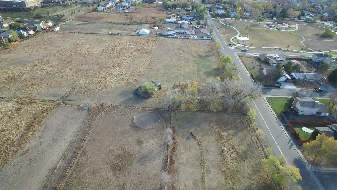 Aerial view in Hidden Lake, Colorado with a broad tan field at left, a residential road at right, tree shadows, houses, and no visible people.