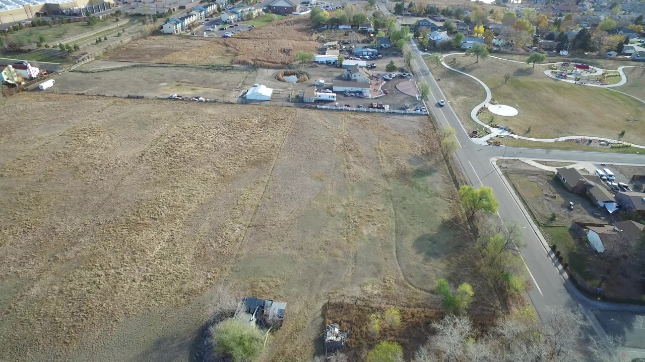 Elevated view in Hidden Lake, Colorado with tan fields across the foreground, a road near the right side, curved white paths, homes, vehicles, and no visible people.