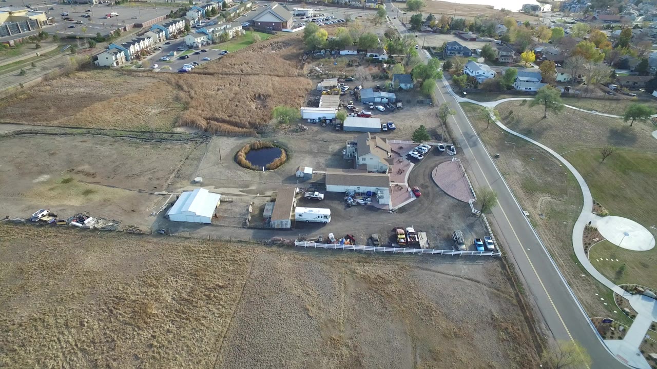 Aerial view of a road beside dry lots, houses, parked vehicles, and a small pond.