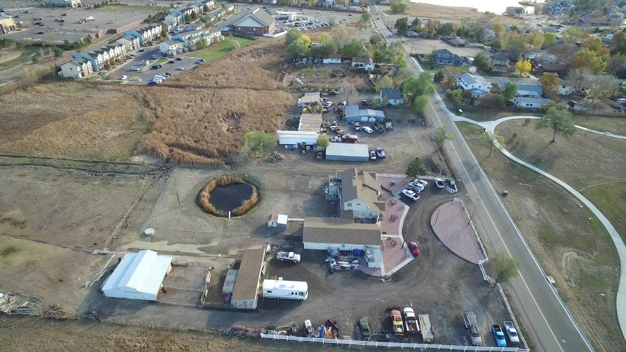 Aerial view of dry fields, a small pond, homes, sheds, trucks, and a roadside property.